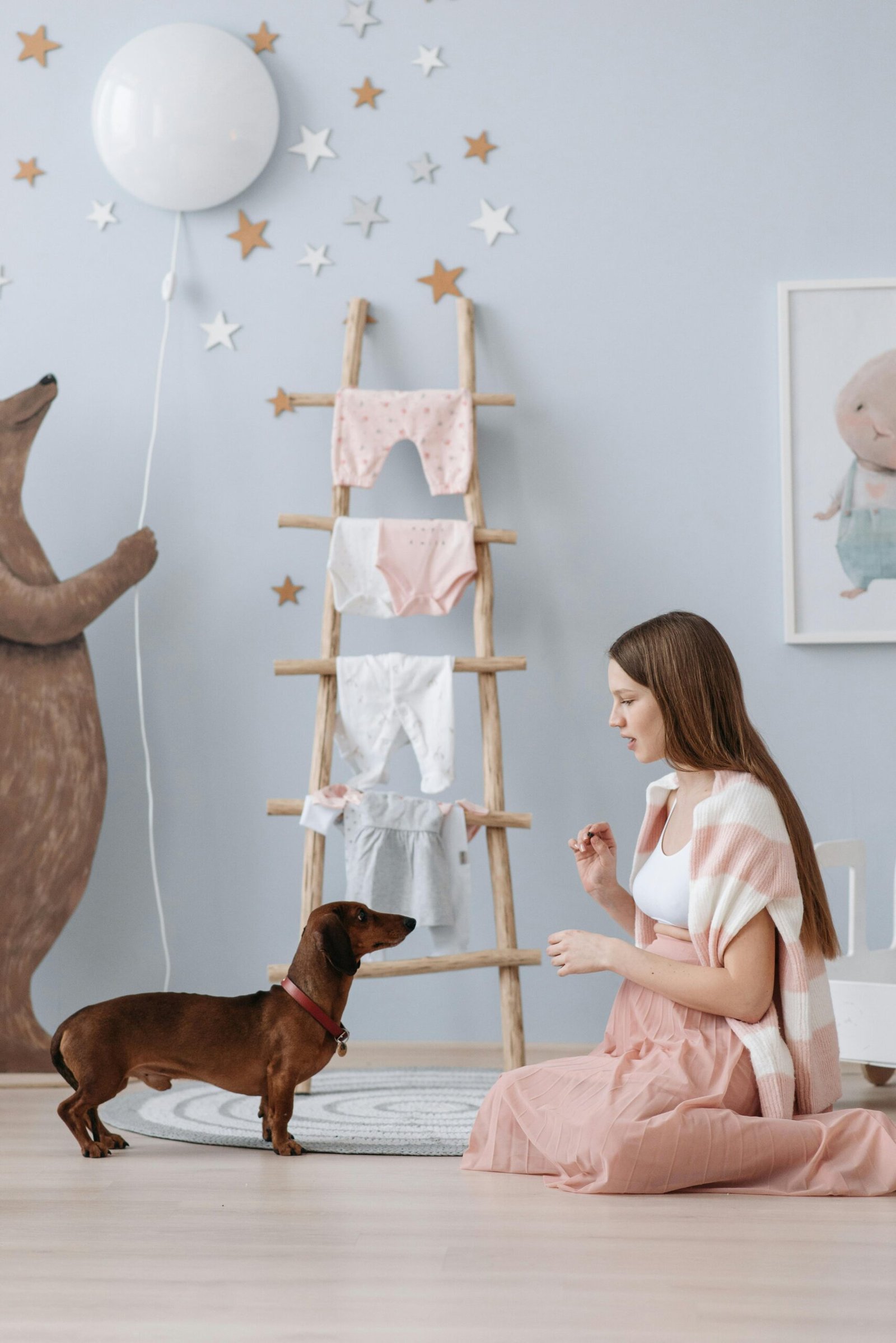 Pregnant woman sits next to a dachshund in a beautifully decorated nursery with baby clothes and star decorations.